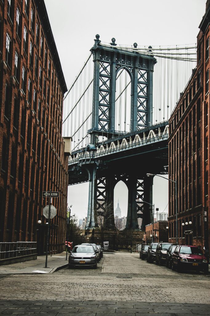 View of Manhattan Bridge between red brick buildings, with cars parked on the street.